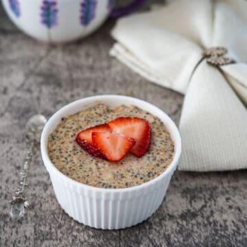 A bowl of chia pudding topped with a sliced strawberry, accompanied by a silver spoon and a cloth napkin, on a wooden table.