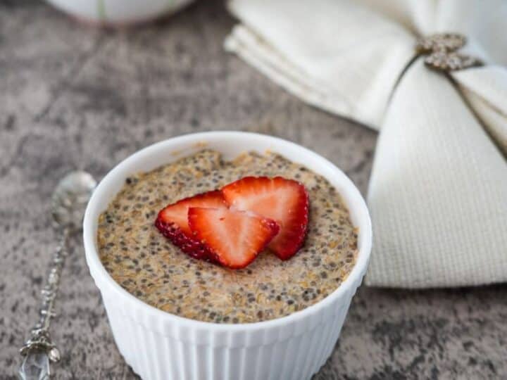 A bowl of chia pudding topped with a sliced strawberry, accompanied by a silver spoon and a cloth napkin, on a wooden table.