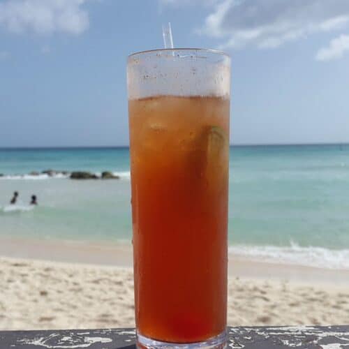 A tall glass of iced tea with a straw sits on a ledge overlooking a sandy beach and the ocean.