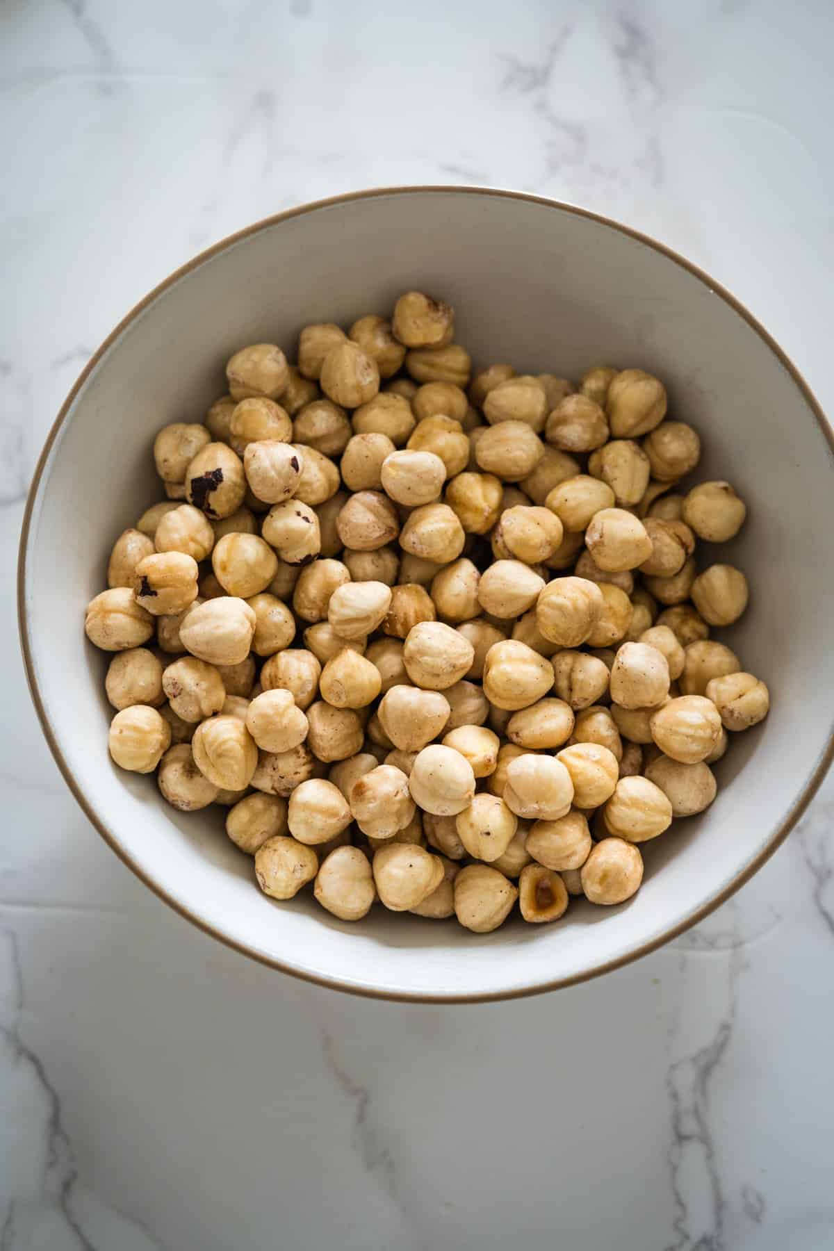 A white bowl filled with hazelnuts, the perfect base for making your own sugar-free Nutella, sits on a marble surface.