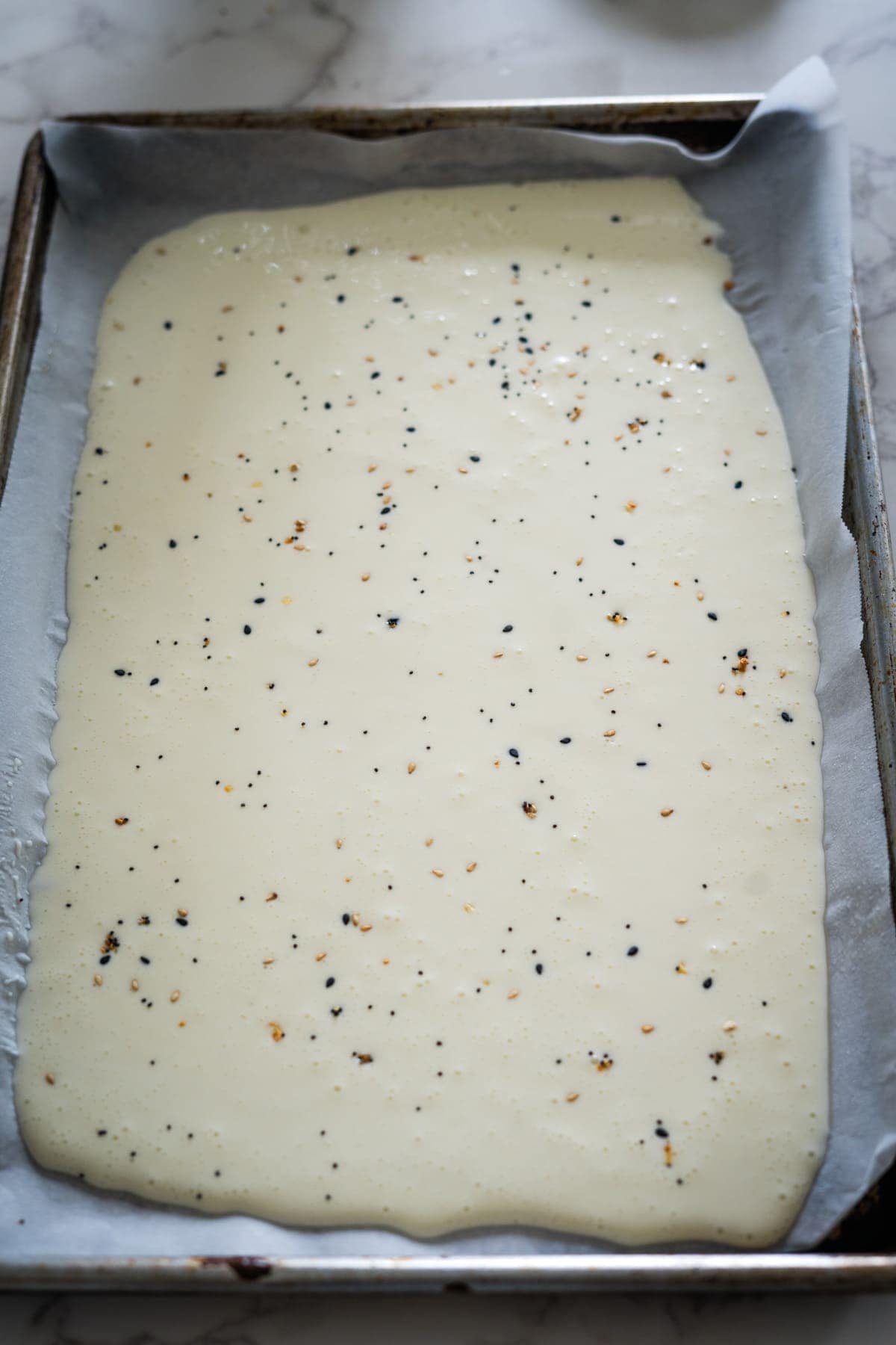 A rectangular baking tray with parchment paper holds a thin, spread-out layer of creamy white batter sprinkled with black and beige specks.