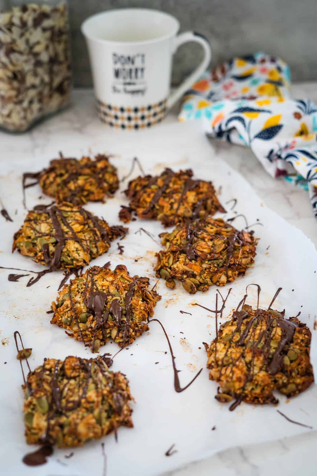 A plate of homemade keto granola cookies drizzled with chocolate, beside a patterned napkin and a mug with text.