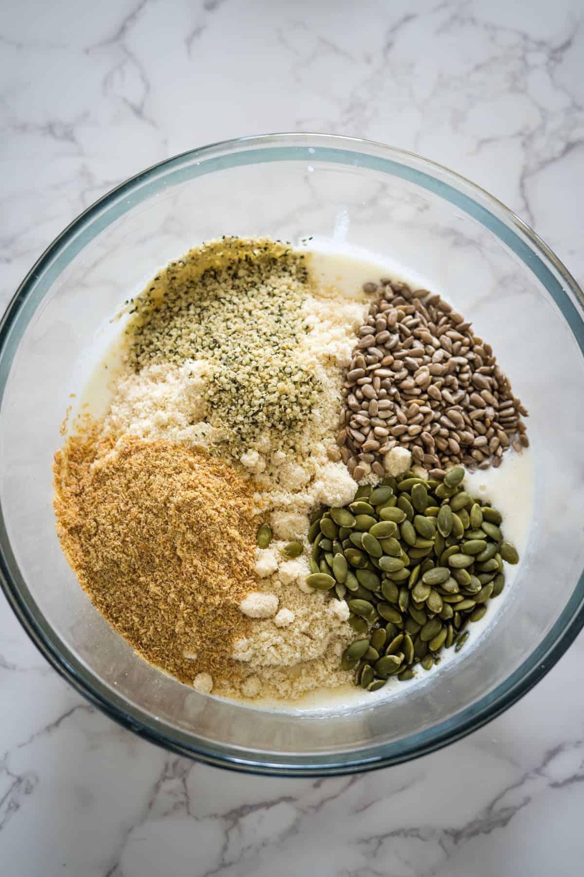 A glass bowl containing a mixture of ingredients, including sunflower seeds, pumpkin seeds, hemp seeds, flaxseed meal, and other powders on a white marble surface.