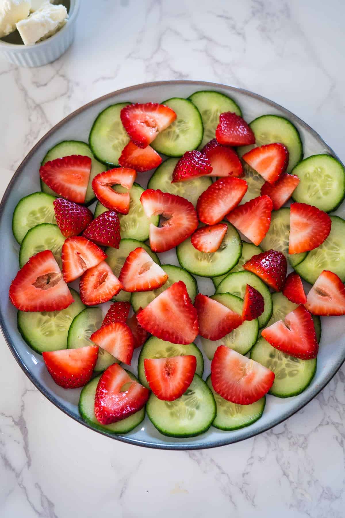 A plate of sliced cucumbers and strawberries arranged in an alternating pattern to form a cucumber strawberry salad on a white marble surface.