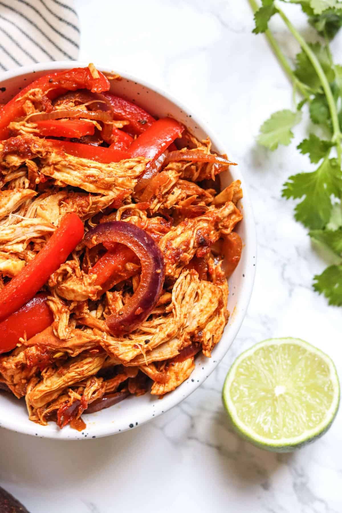 A bowl of shredded chicken with red peppers and onions, garnished with a lime wedge and cilantro, on a marble surface.