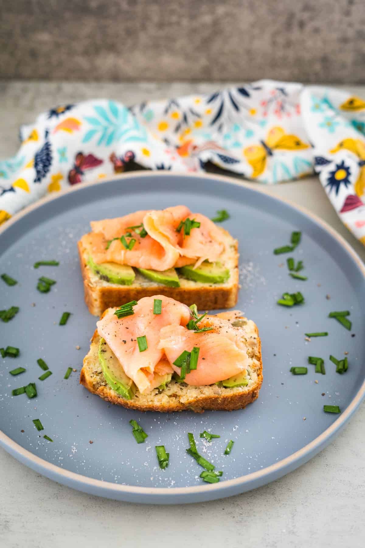 A gray plate with two slices of toast topped with avocado, smoked salmon, and fresh chives. A colorful cloth is in the background, complementing the vibrant display on the cottage cheese bread.