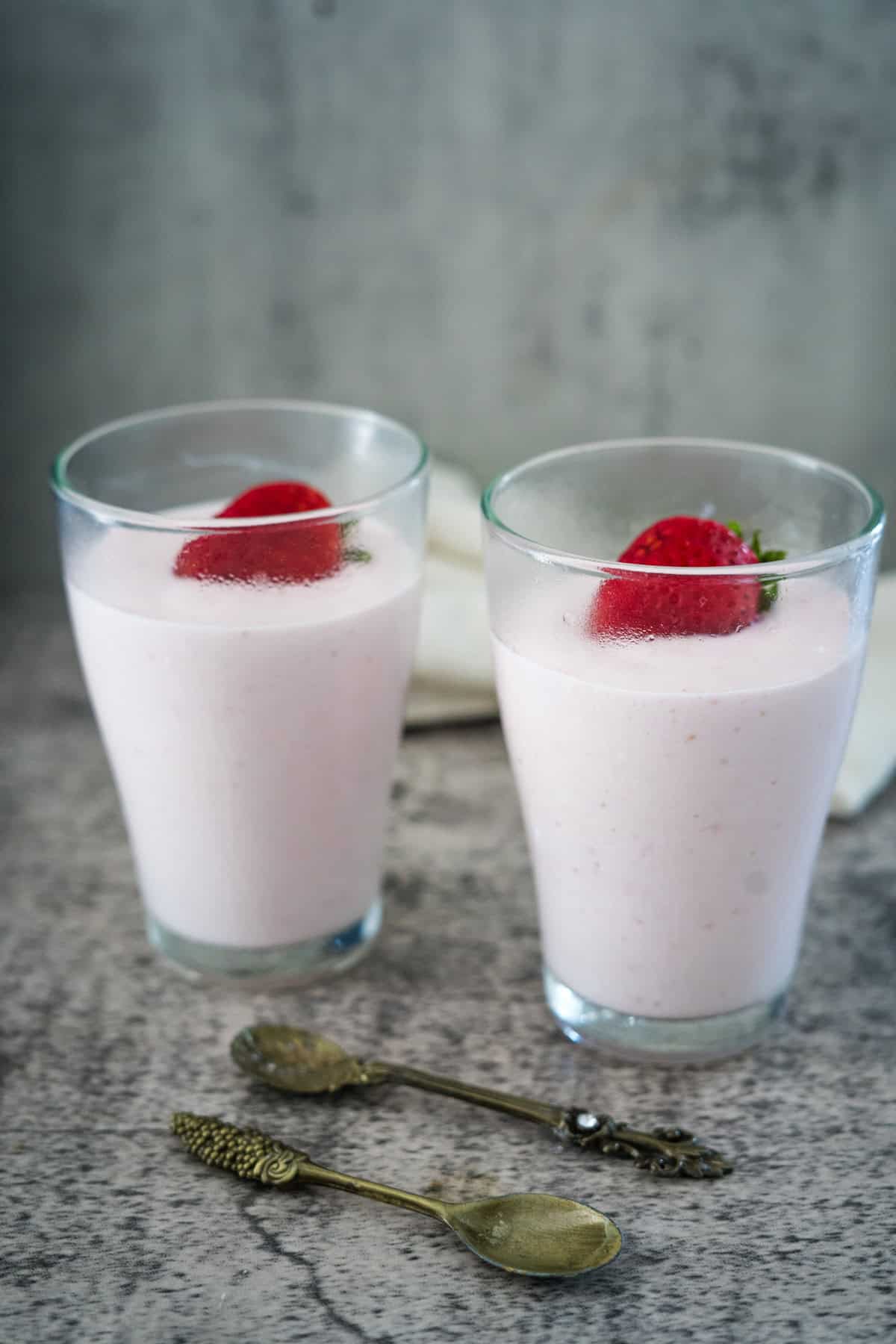 Two glasses filled with pink strawberry smoothies, each topped with a whole strawberry, are placed on a textured surface with two vintage-style spoons in front.