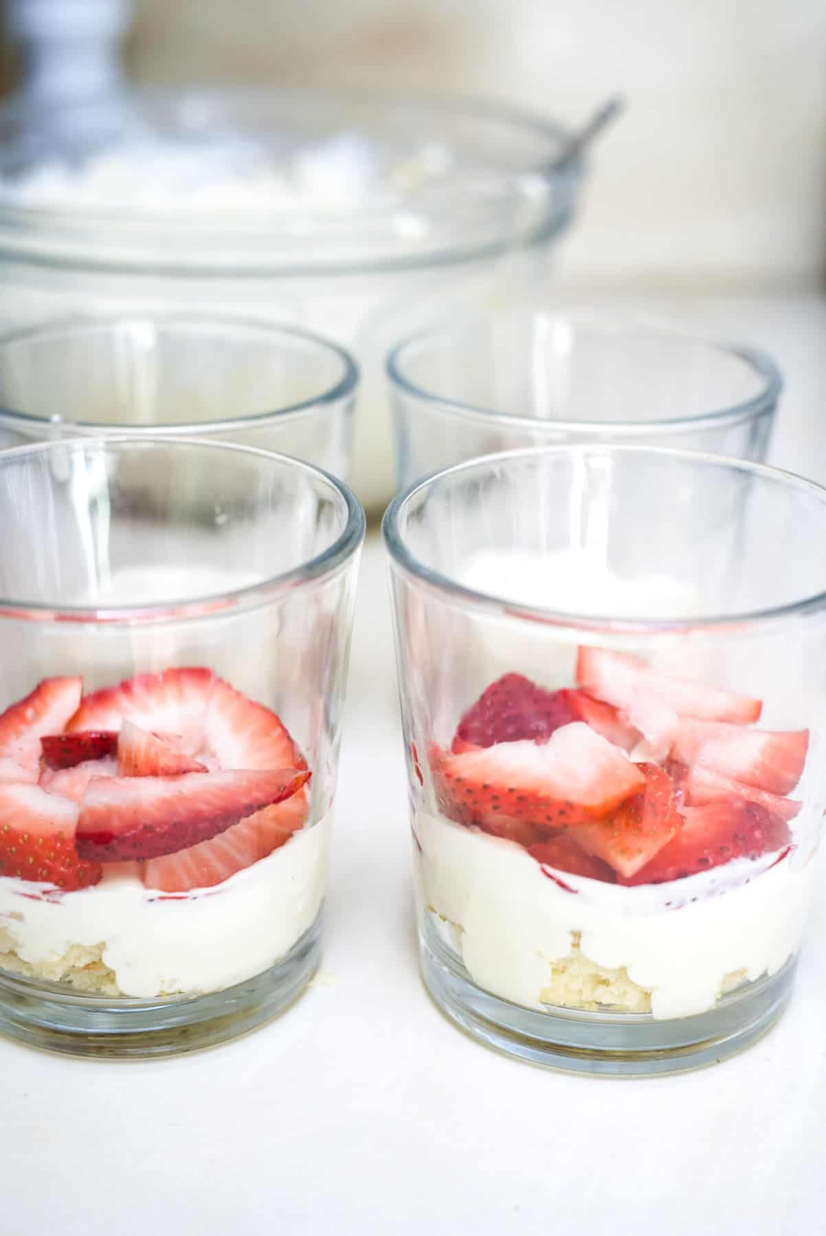 Four glass cups containing a dessert with layers of cream and sliced strawberries. A large bowl with more cream is visible in the background.