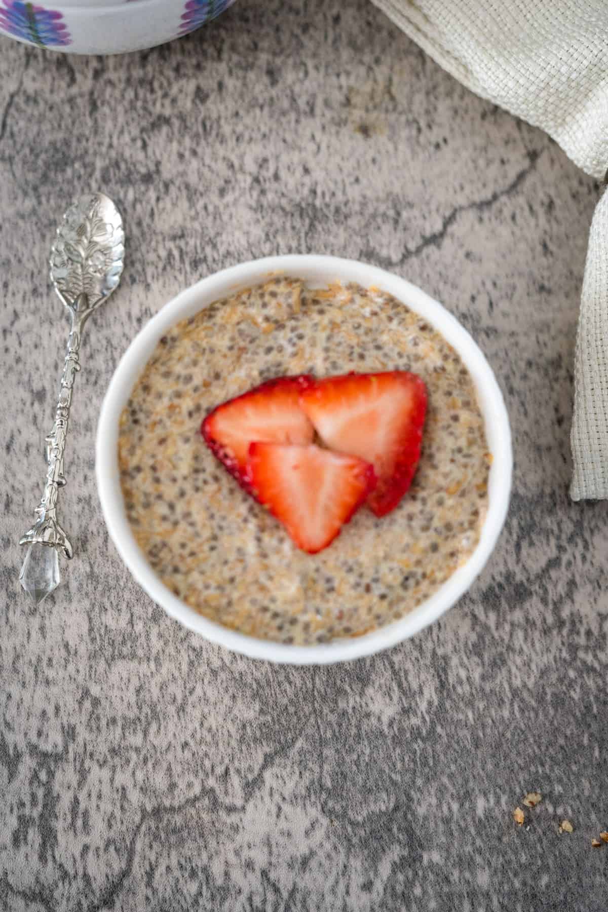 A bowl of chia and flaxseed pudding topped with a sliced strawberry, next to a decorative spoon on a textured grey surface.