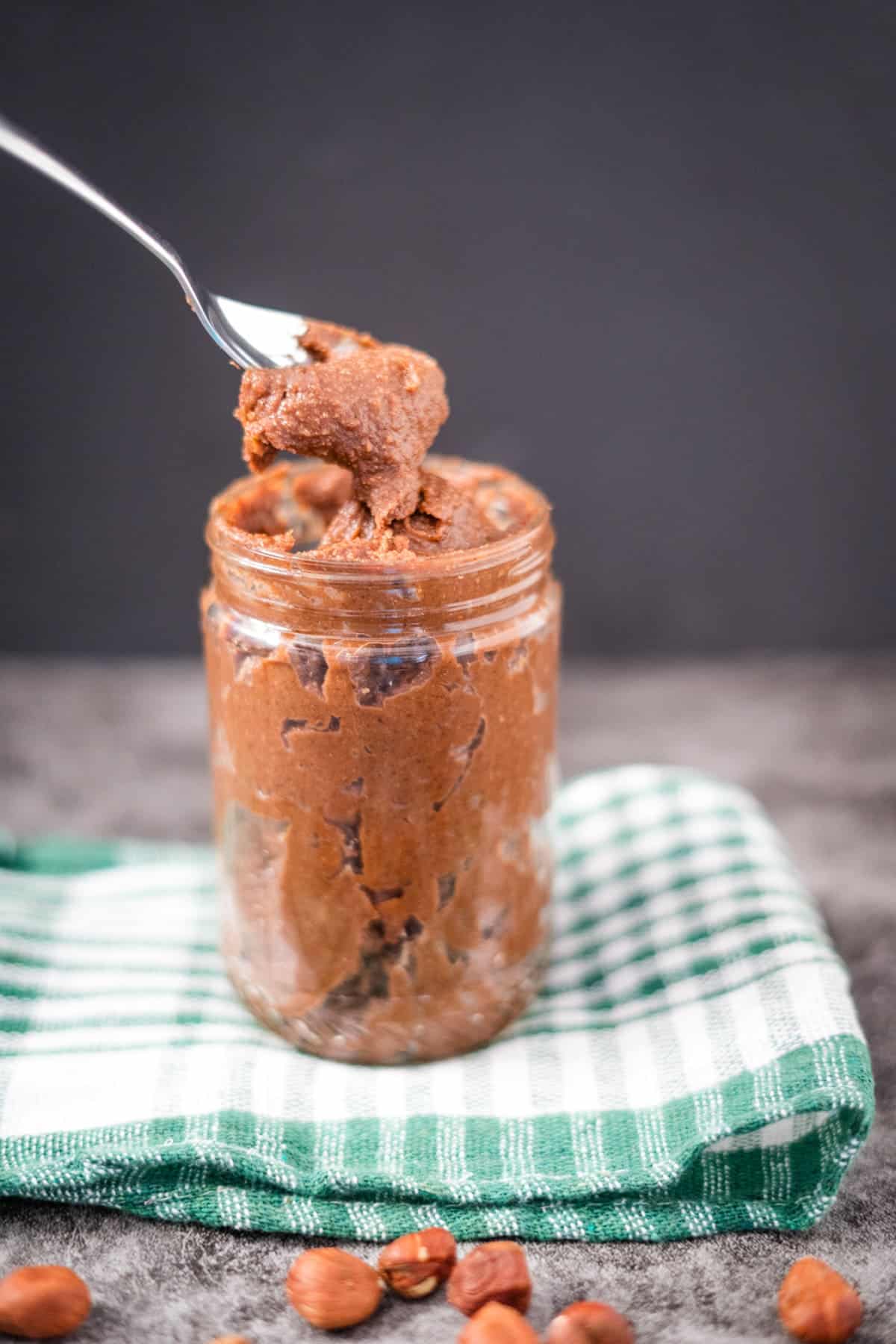 A spoon scoops out chocolate hazelnut spread from a glass jar resting on a green and white checkered cloth, with hazelnuts scattered nearby.