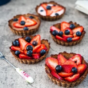 Five small tarts topped with sliced strawberries and blueberries are arranged on a gray surface next to a fork with a floral handle.