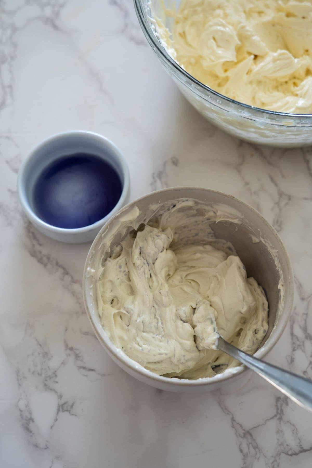 A small bowl filled with whipped cream, another bowl with a blue liquid, and a larger bowl containing more whipped cream sit on a marble countertop, perfectly complementing the lemon lavender cupcakes nearby.