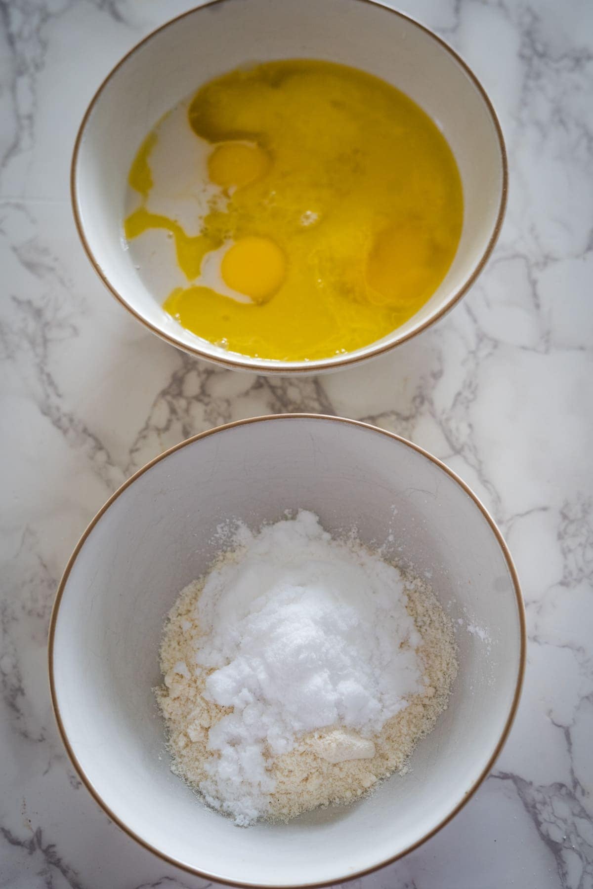 Two mixing bowls on a marble counter: the top bowl contains eggs and a yellow liquid, hinting at zesty lemon. The bottom bowl holds a mix of flour, sugar, and baking powder, all coming together for delicious lemon lavender cupcakes.