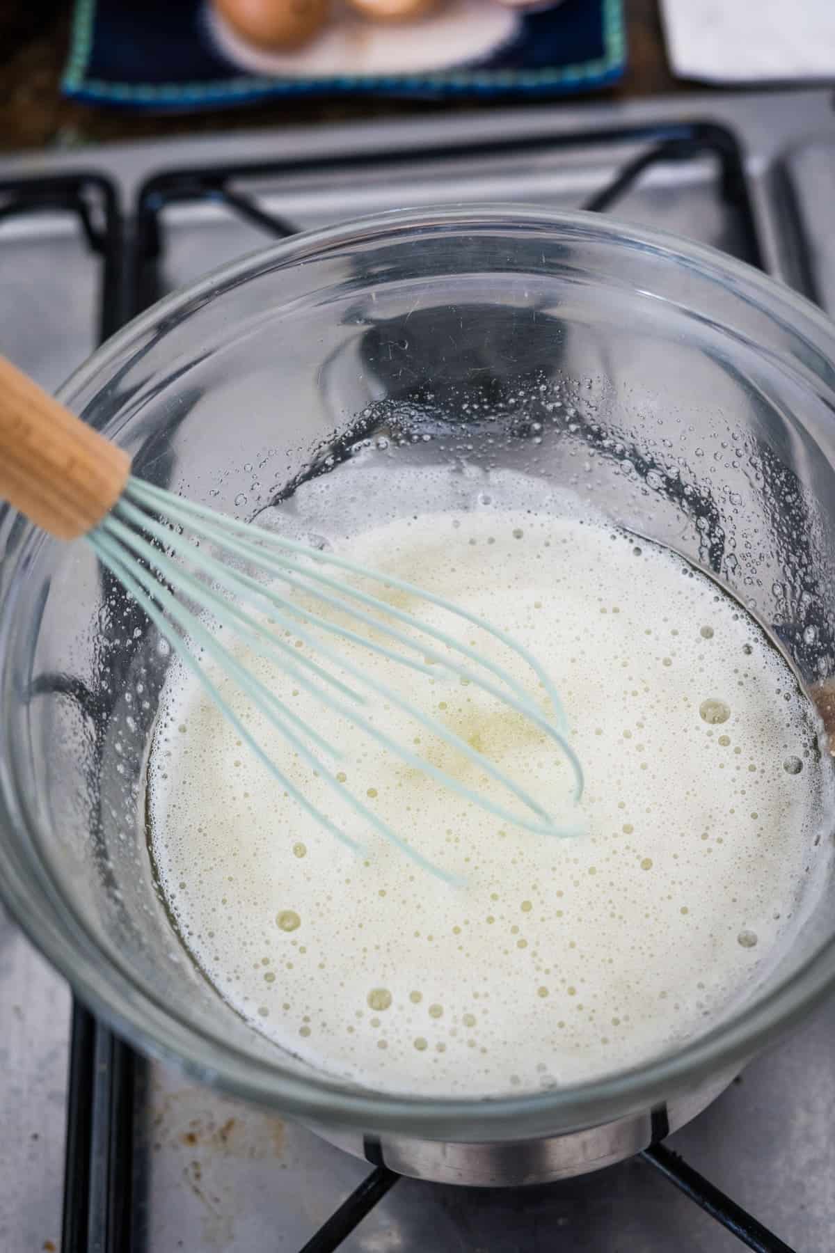 A whisk is mixing foamy liquid in a glass bowl on a stovetop, preparing the base for delicious lemon lavender cupcakes.