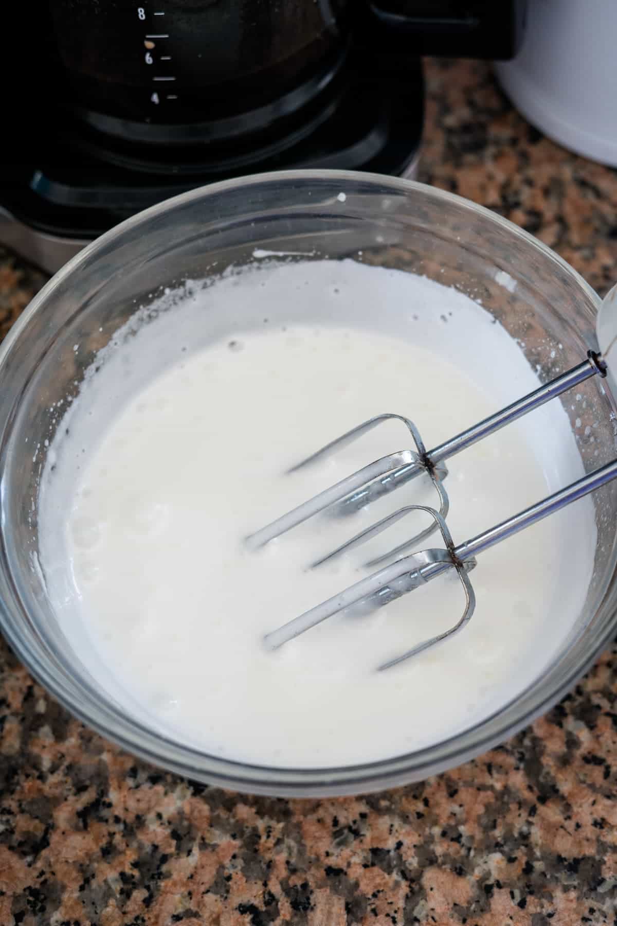 A glass bowl of white liquid is being mixed with an electric hand mixer on a granite countertop, perhaps the start of delicious lemon lavender cupcakes. A coffee maker is visible in the background.