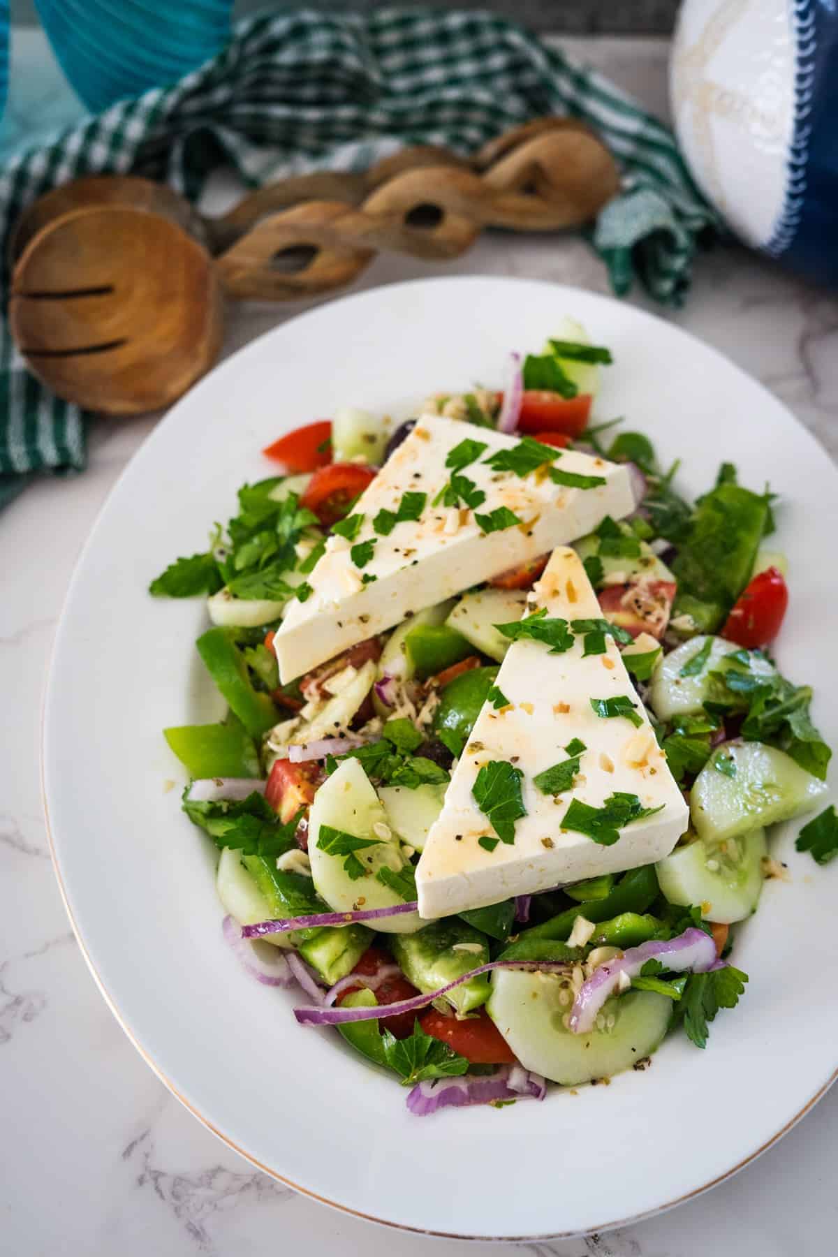 A Greek village salad with cucumbers, tomatoes, red onions, green peppers, and parsley, topped with two large slices of cheese. It's served on a white plate with wooden utensils and a green checkered cloth in the background.