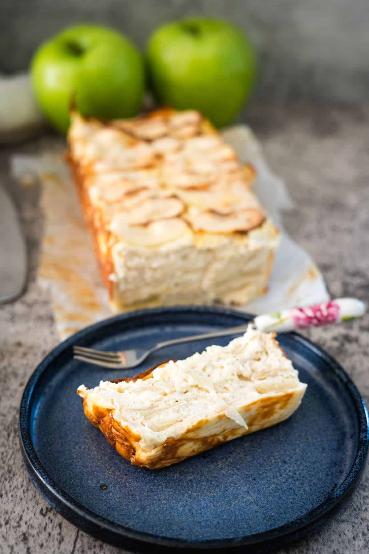 A slice of layered apple cream cheese cake on a blue plate, with the full dessert in the background accompanied by two green apples. A fork rests on the plate next to the dessert slice.