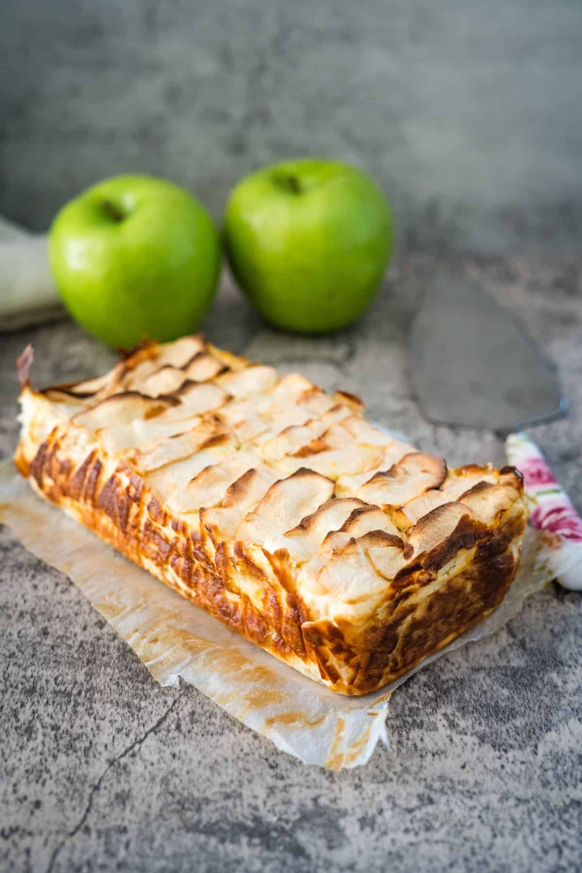 A rectangular apple cream cheese cake topped with thin apple slices is placed on parchment paper. Two fresh green apples and a pastry server are in the background.