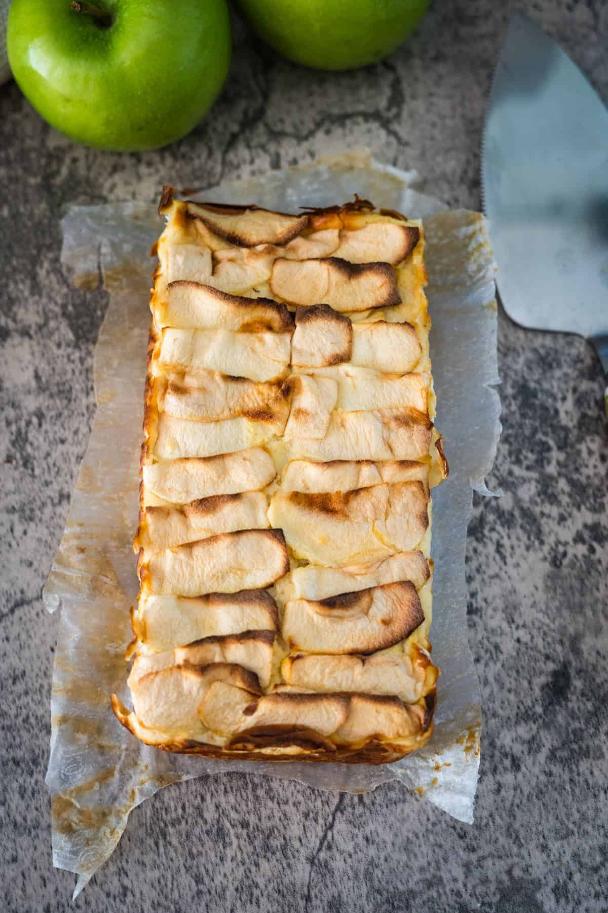 A rectangular apple tart with a browned top sits on parchment paper next to a green apple and a dessert server. The background is a textured stone surface.