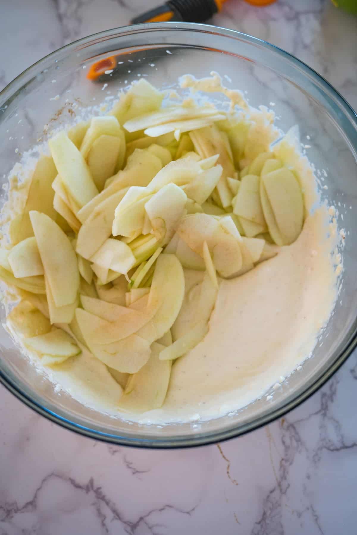 A glass bowl contains sliced apples mixed with a creamy batter on a marble countertop.