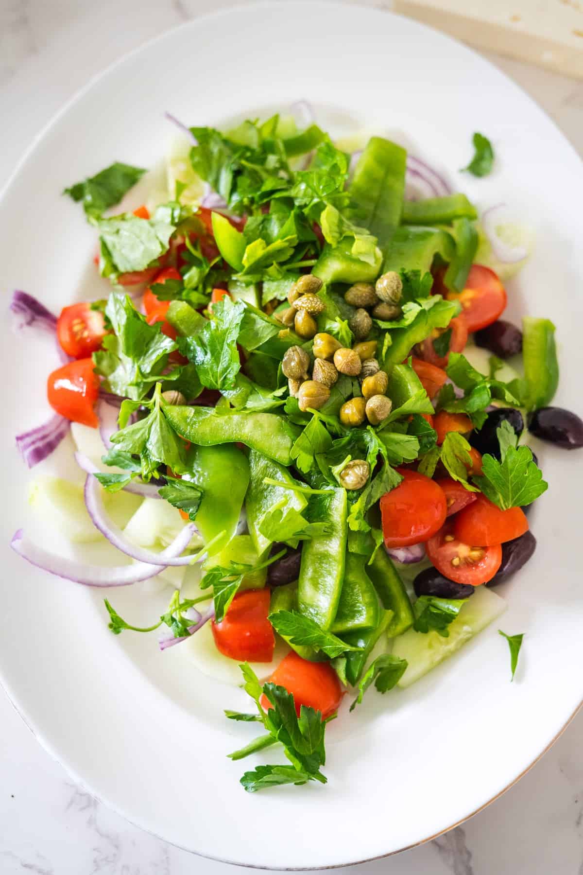 A Greek village salad with cherry tomatoes, snow peas, red onion, parsley, capers, and olives on a white plate.