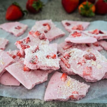 Pieces of pink strawberry yogurt bark topped with shredded coconut and small strawberry chunks on parchment paper with whole strawberries in the background.