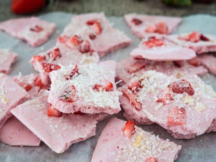 Pieces of pink strawberry yogurt bark topped with shredded coconut and small strawberry chunks on parchment paper with whole strawberries in the background.