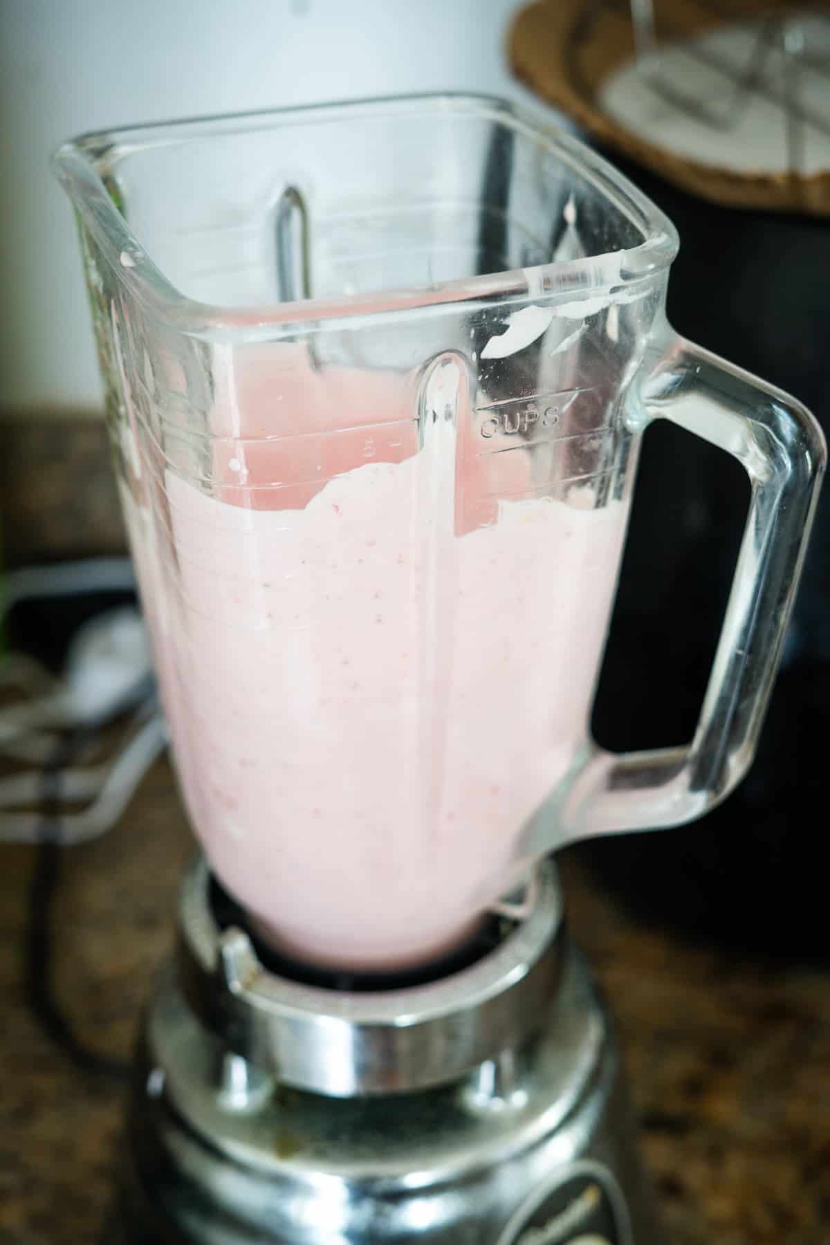 A glass blender container filled with a pink smoothie mixture sits on a kitchen countertop, next to pieces of frozen cottage cheese bark.