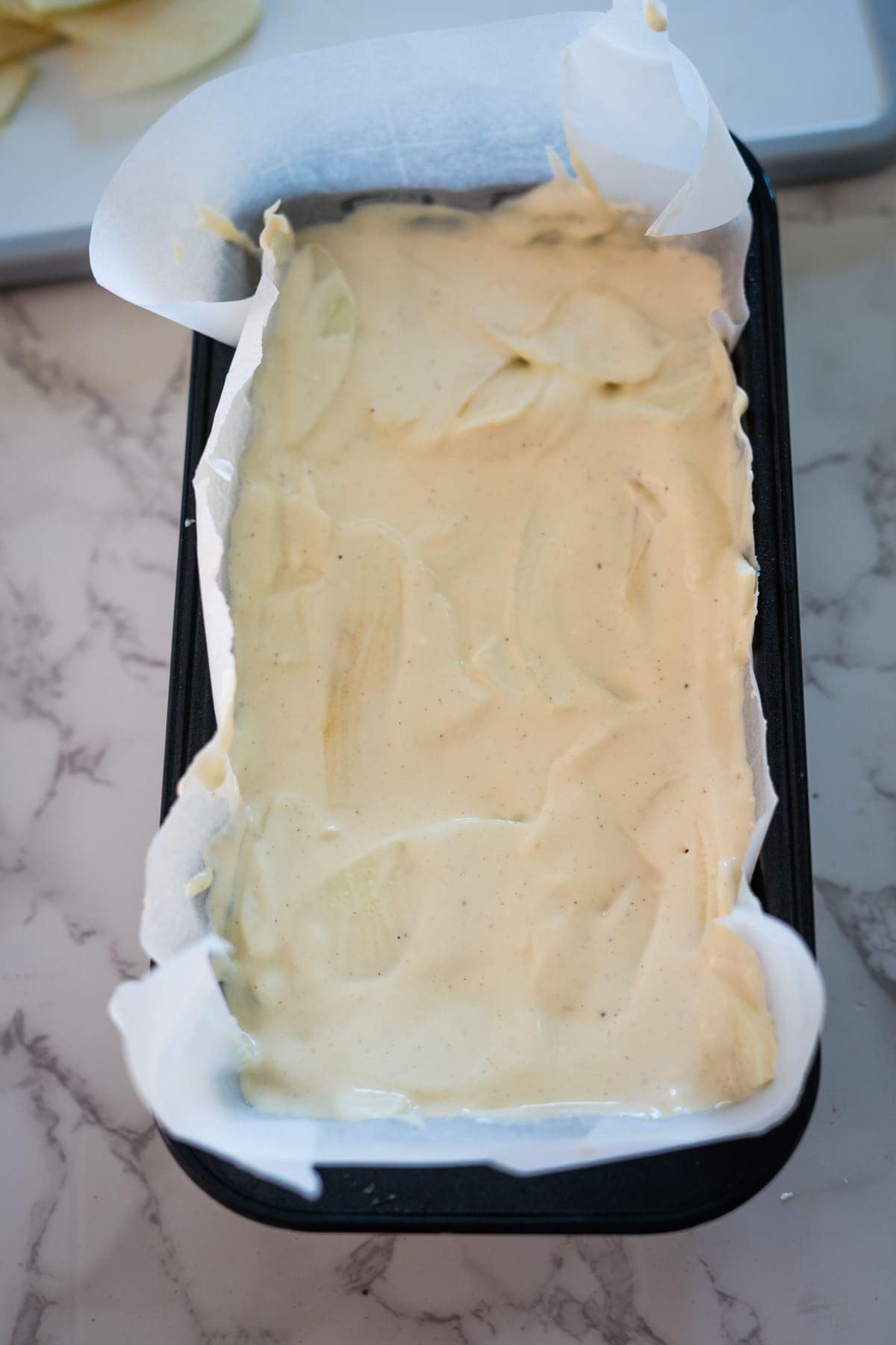 A rectangular baking pan lined with parchment paper, filled with a creamy batter, sits on a marbled countertop.