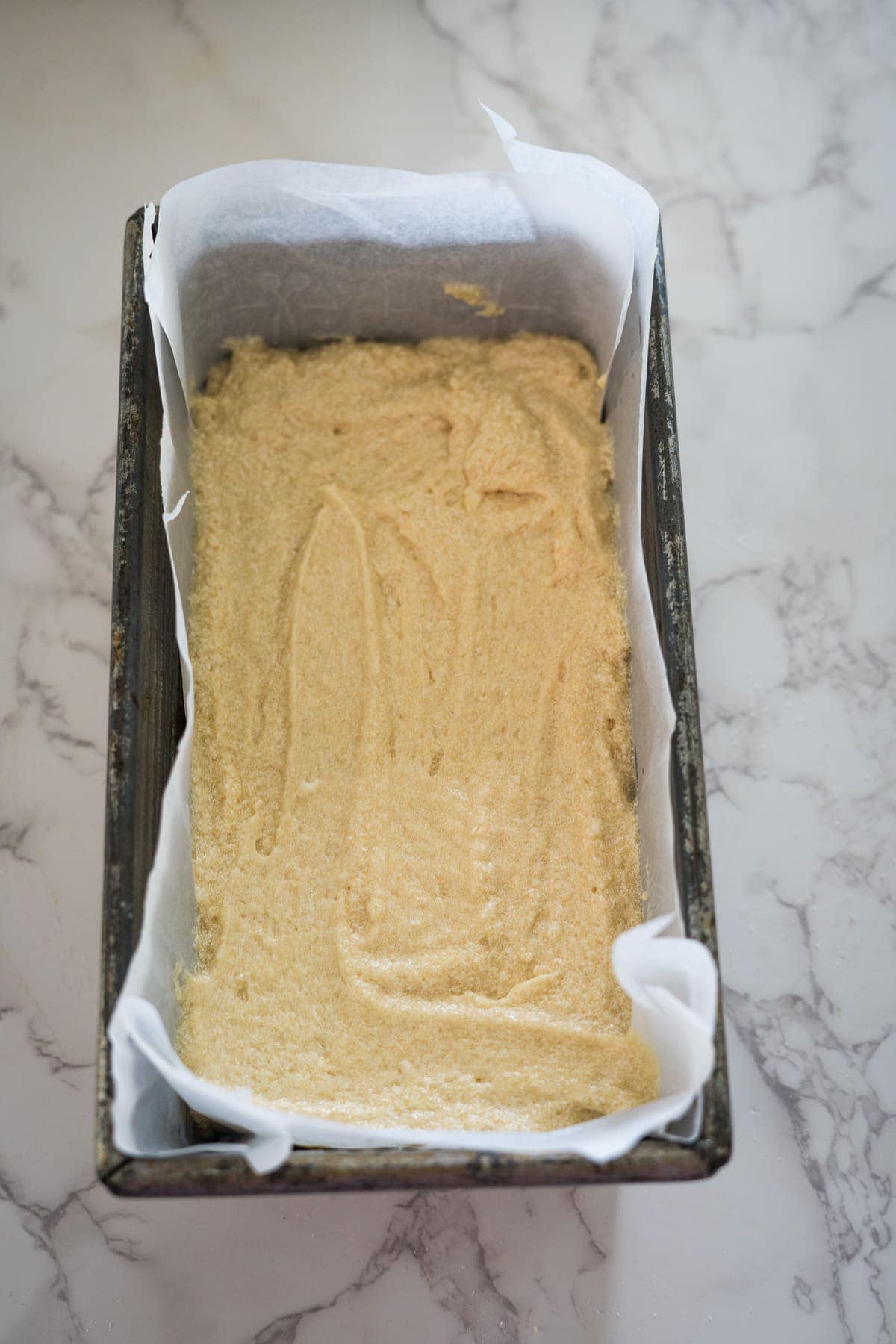 A loaf pan lined with parchment paper is filled with raw cake batter, placed on a marble countertop.