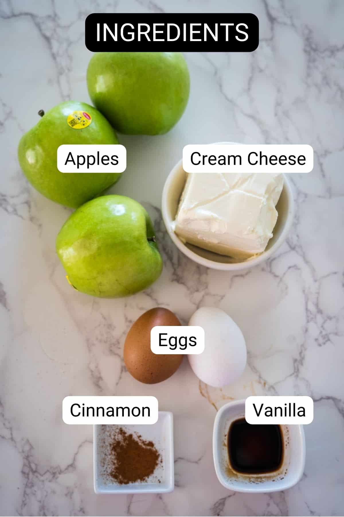 Image showing labeled ingredients on a marble counter: three green apples, a block of cream cheese, two eggs, a small amount of cinnamon, and vanilla extract.