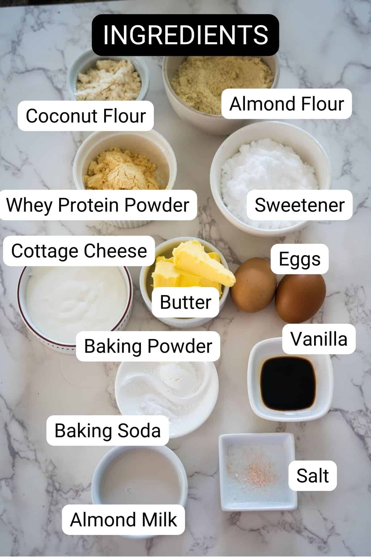 A collection of labeled baking ingredients is arranged on a marble countertop, including almond flour, coconut flour, whey protein powder, sweetener, cottage cheese, butter, eggs, vanilla, and various other items.