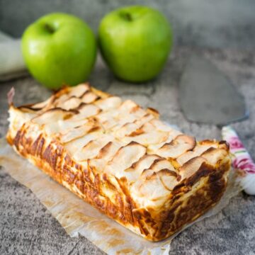 Rectangular apple cream cheese cake with browned edges, placed on parchment paper. Two green apples and a cake server are in the background.