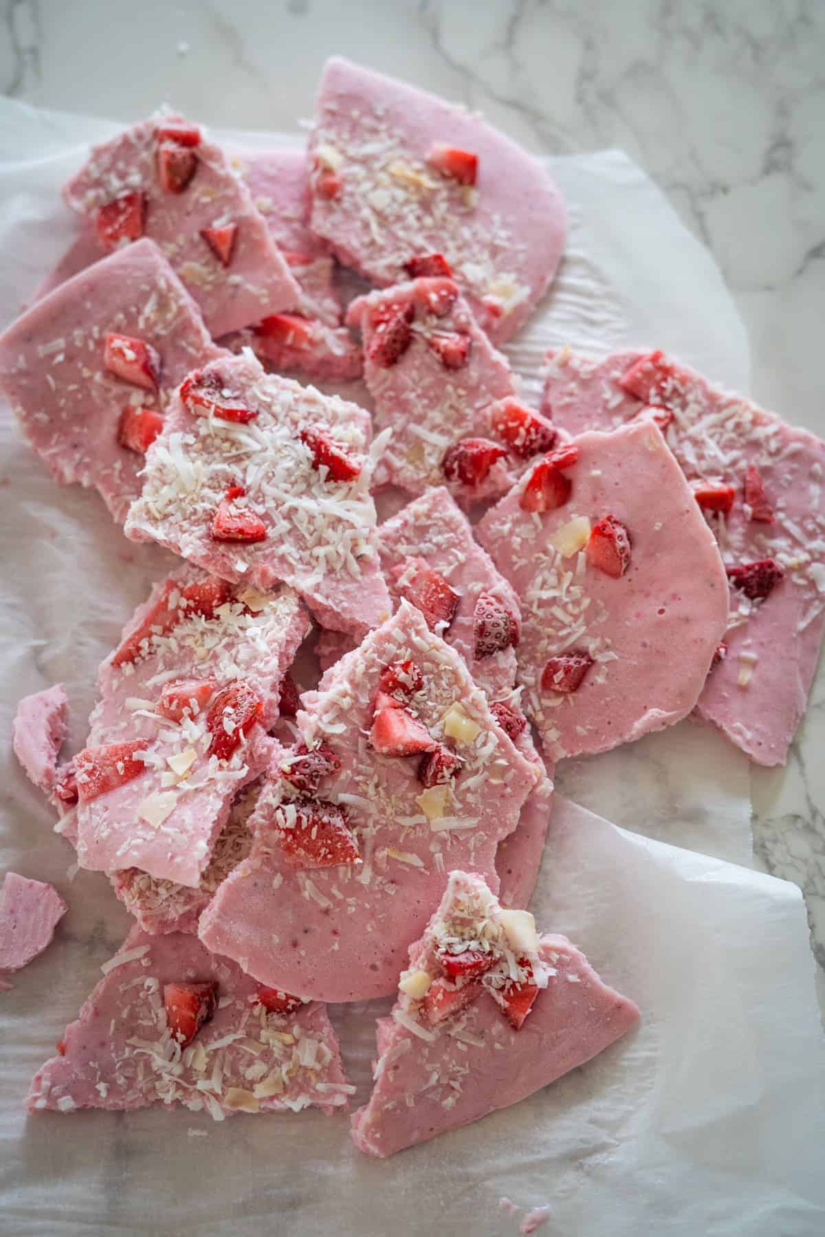 Broken pieces of frozen cottage cheese bark are topped with sliced strawberries and shredded coconut, arranged on parchment paper.