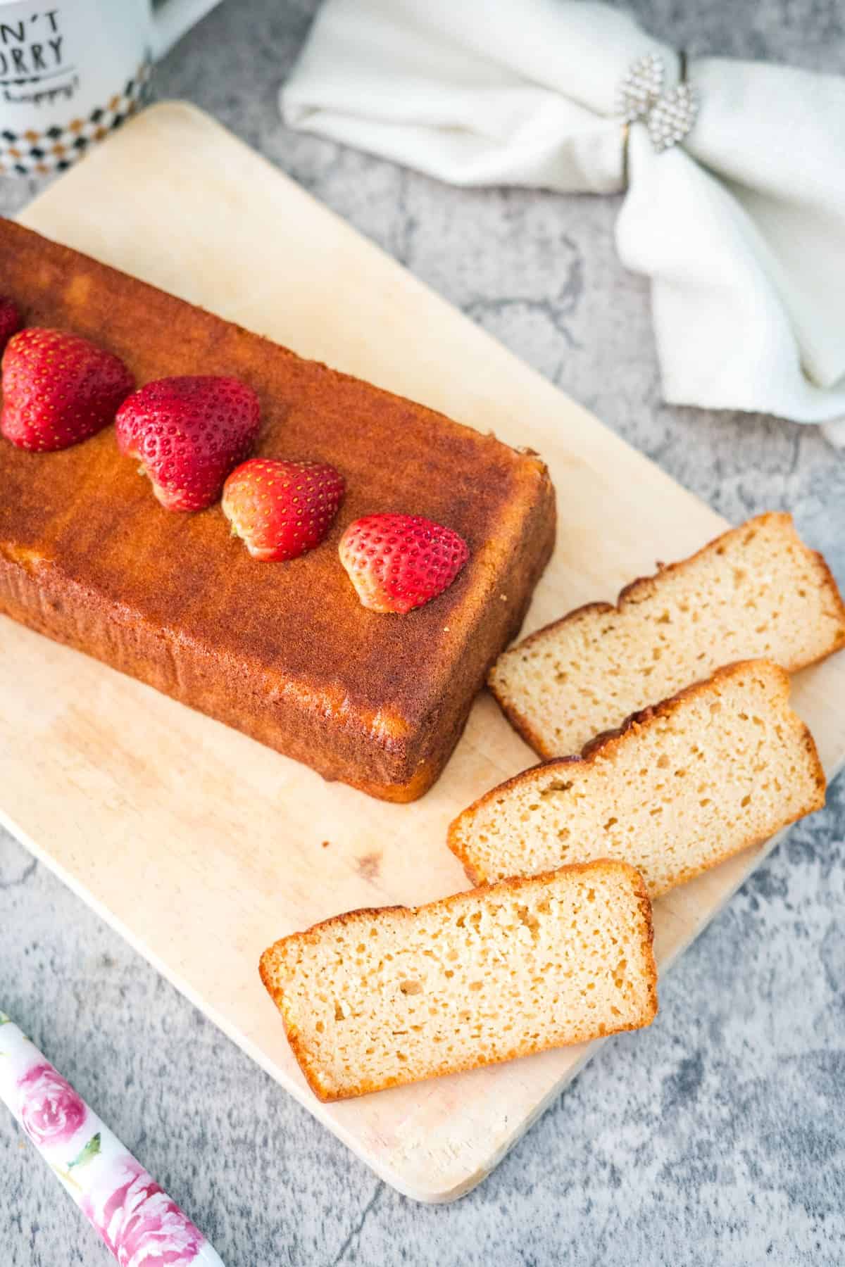 A rectangular loaf cake sits on a wooden cutting board, topped with fresh strawberries. Three slices of the cottage cheese pound cake are cut and placed next to the loaf. A floral knife and a napkin are in the background.