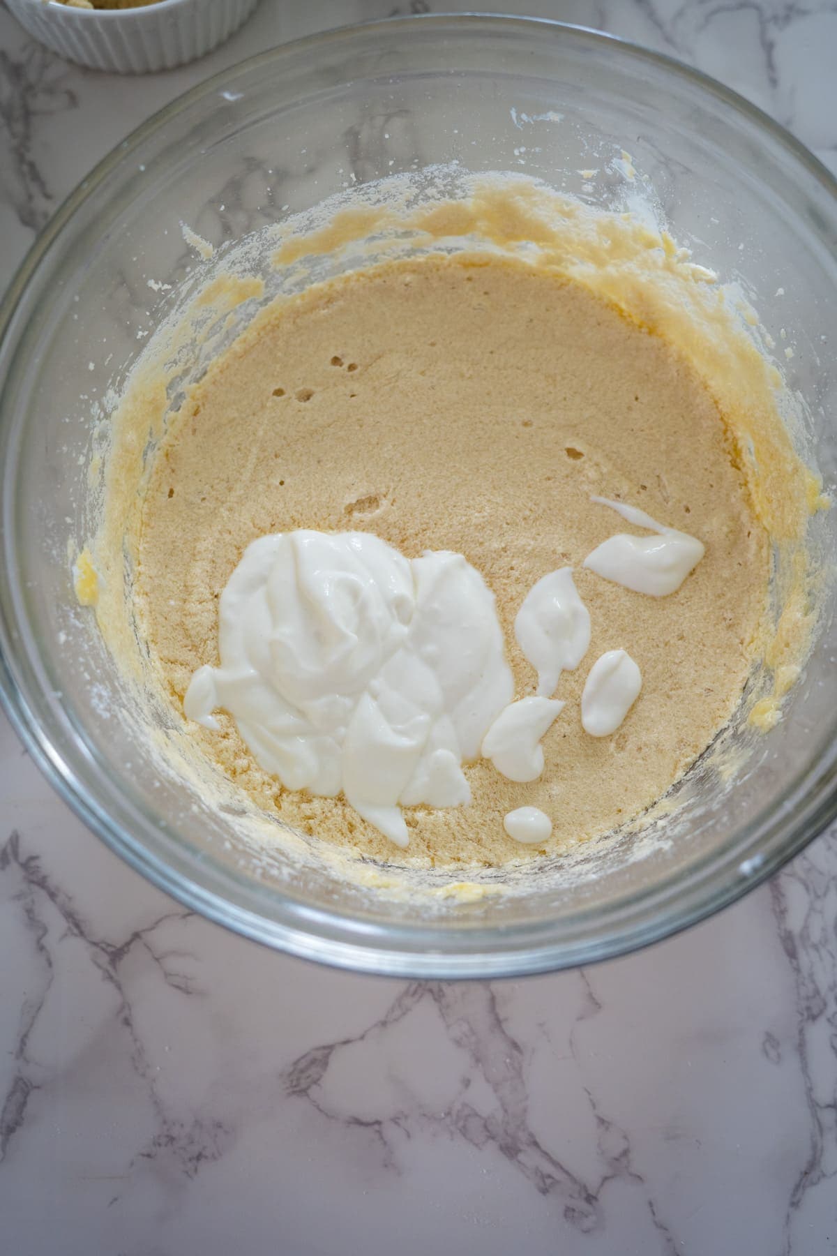 A glass bowl on a marble surface contains a partially mixed dough with a dollop of yogurt on top.