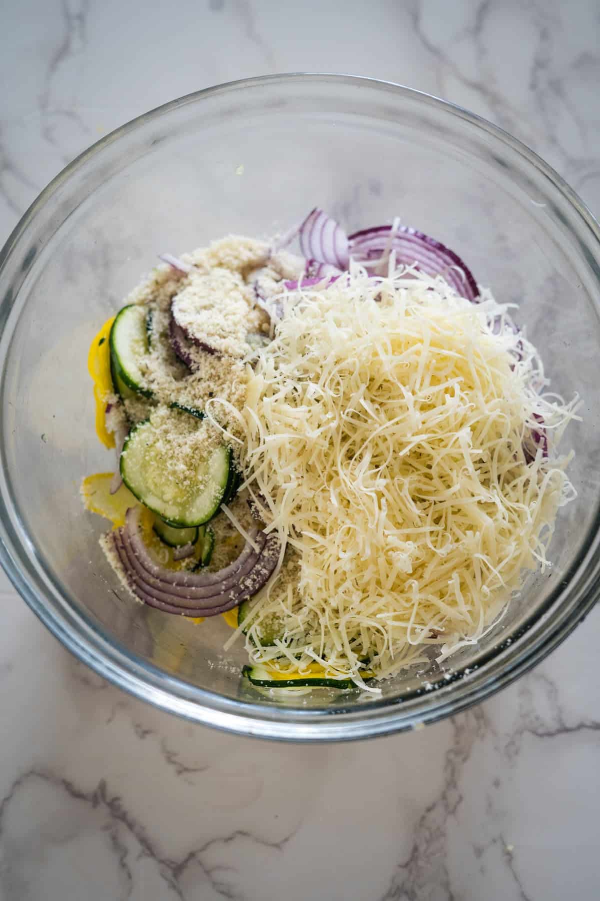 A glass mixing bowl contains sliced zucchini, red onions, grated cheese, and breadcrumbs on a marble countertop.