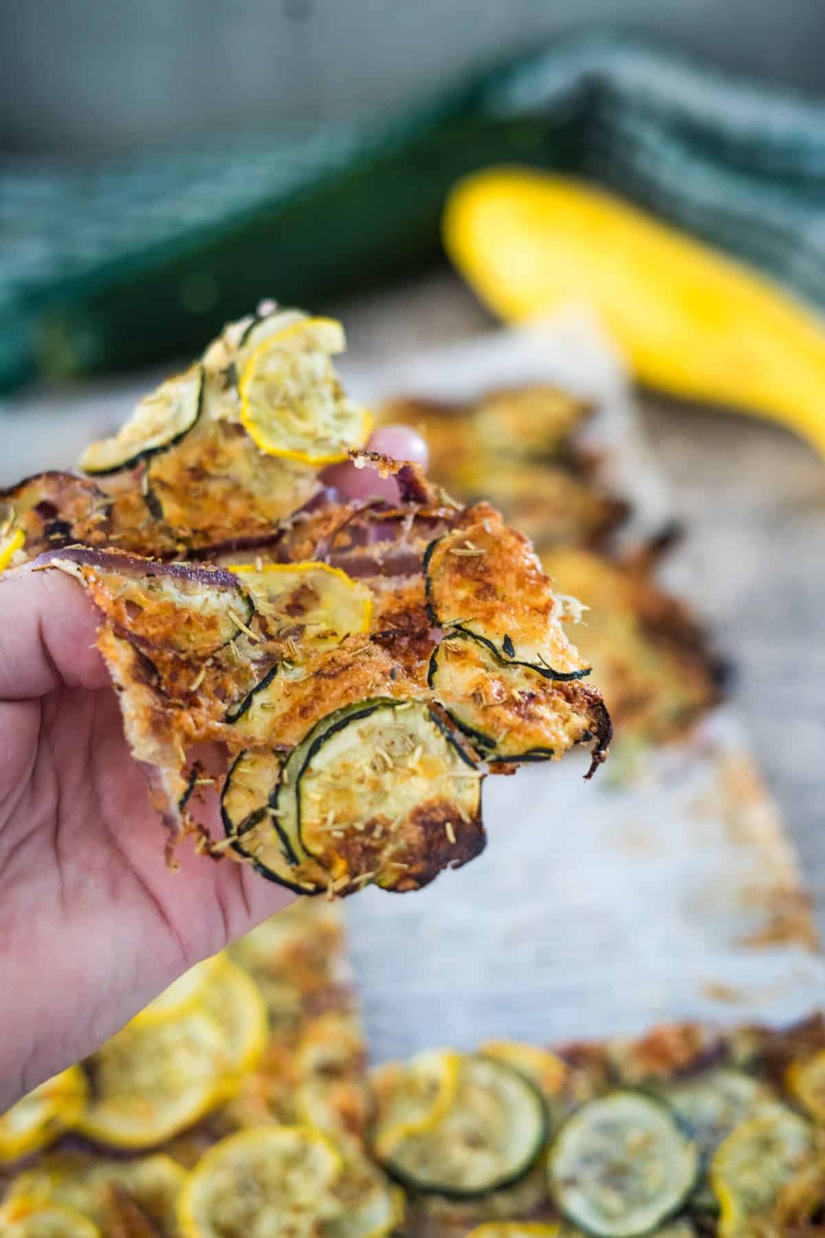 A hand holding a crisp zucchini chip with visible yellow and green zucchini slices in front of a tray of more zucchini chips. A yellow squash and a cucumber are blurred in the background.