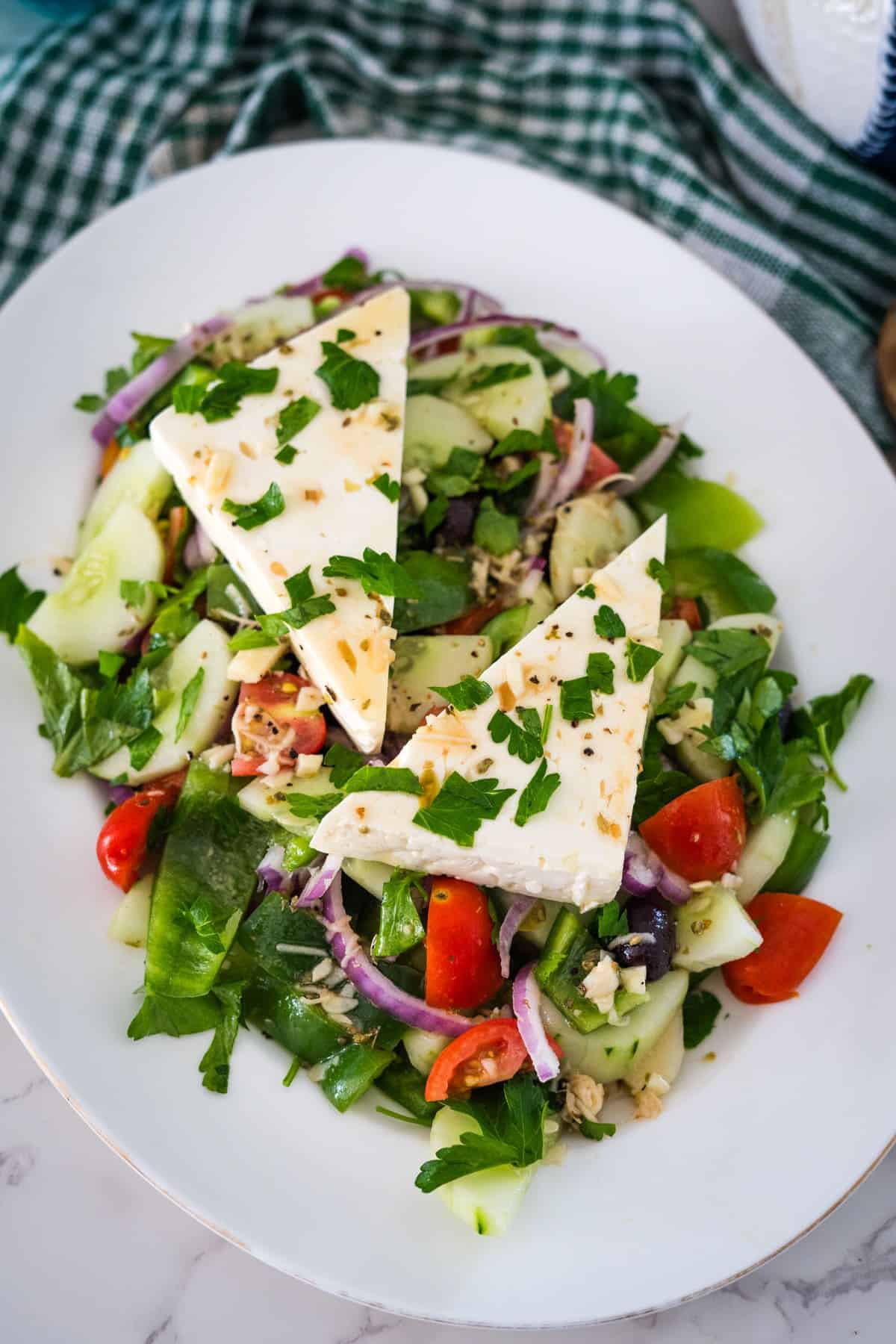 A plate of Greek salad with sliced cucumbers, tomatoes, red onions, and green peppers, topped with two triangular pieces of feta cheese and garnished with parsley.