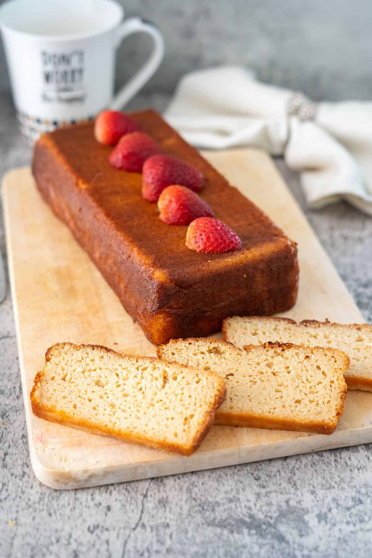 A rectangular loaf cake topped with halved strawberries is placed on a wooden cutting board. Several slices of the cake are cut and laid out on the board beside the loaf. A coffee cup and napkin are in the background.