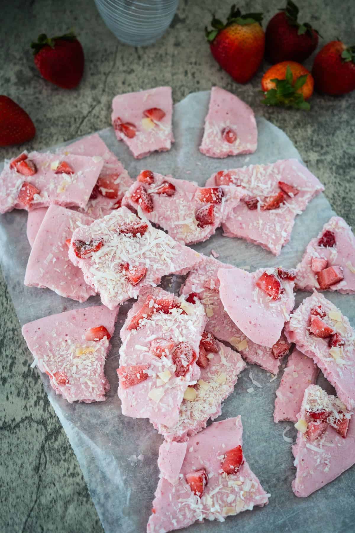 Pink yogurt bark with pieces of strawberry and shaved coconut is arranged on a piece of parchment paper. Some fresh strawberries are in the background, adding a touch of vibrant color. This delightful frozen cottage cheese bark is a refreshing treat perfect for any occasion.