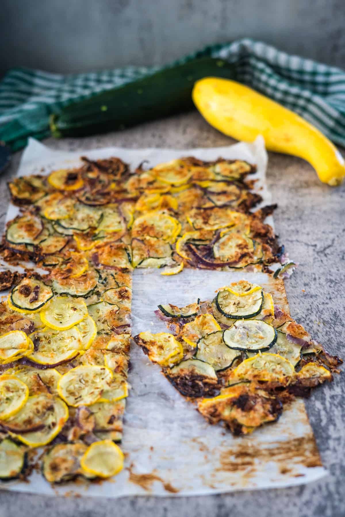 A baked zucchini scarpaccia and yellow squash flatbread on parchment paper with a slice removed. There is a green and white checkered cloth and fresh zucchini and yellow squash in the background.