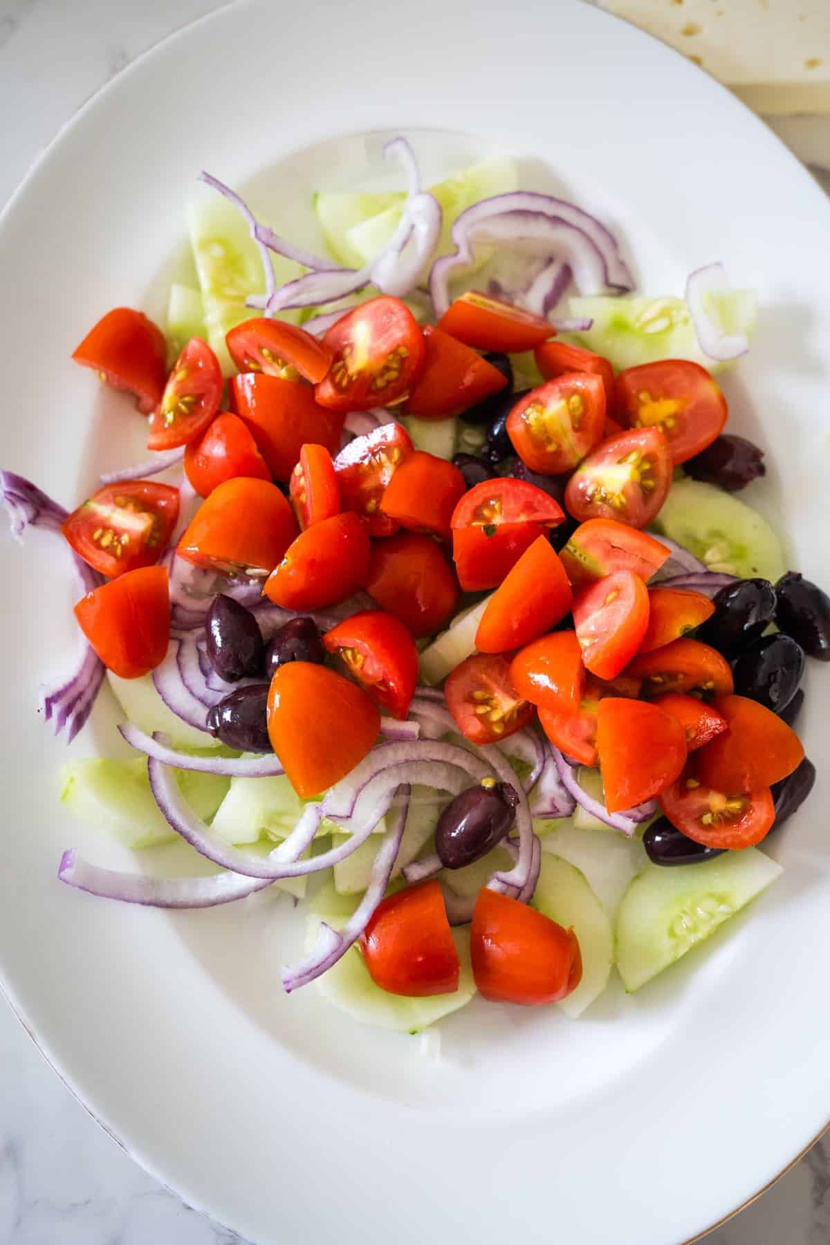 A white plate holds a Greek village salad featuring fresh chopped cherry tomatoes, sliced cucumbers, red onion rings, and black olives.