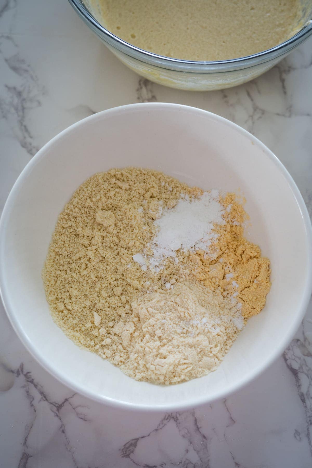 A white bowl containing dry baking ingredients, including brown sugar, white sugar, and flour, on a marble surface.