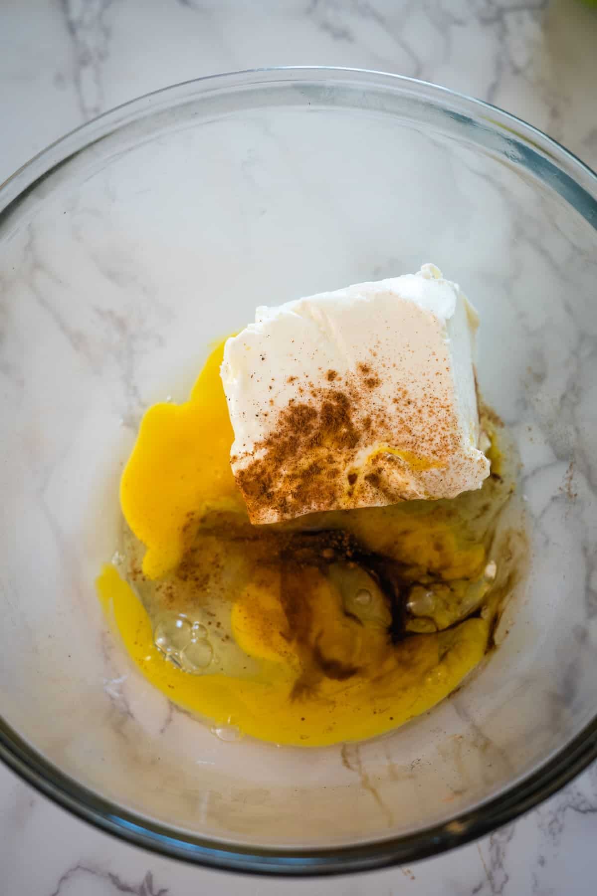 A glass bowl containing a block of cream cheese, a raw egg, vanilla extract, and a pinch of cinnamon on a marble surface.