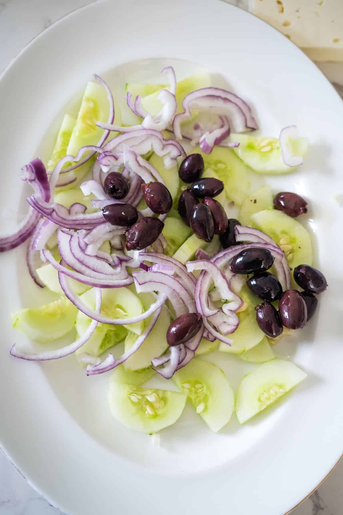 A white plate with sliced cucumbers, red onion, and black olives arranged on it, reminiscent of a classic Greek village salad.