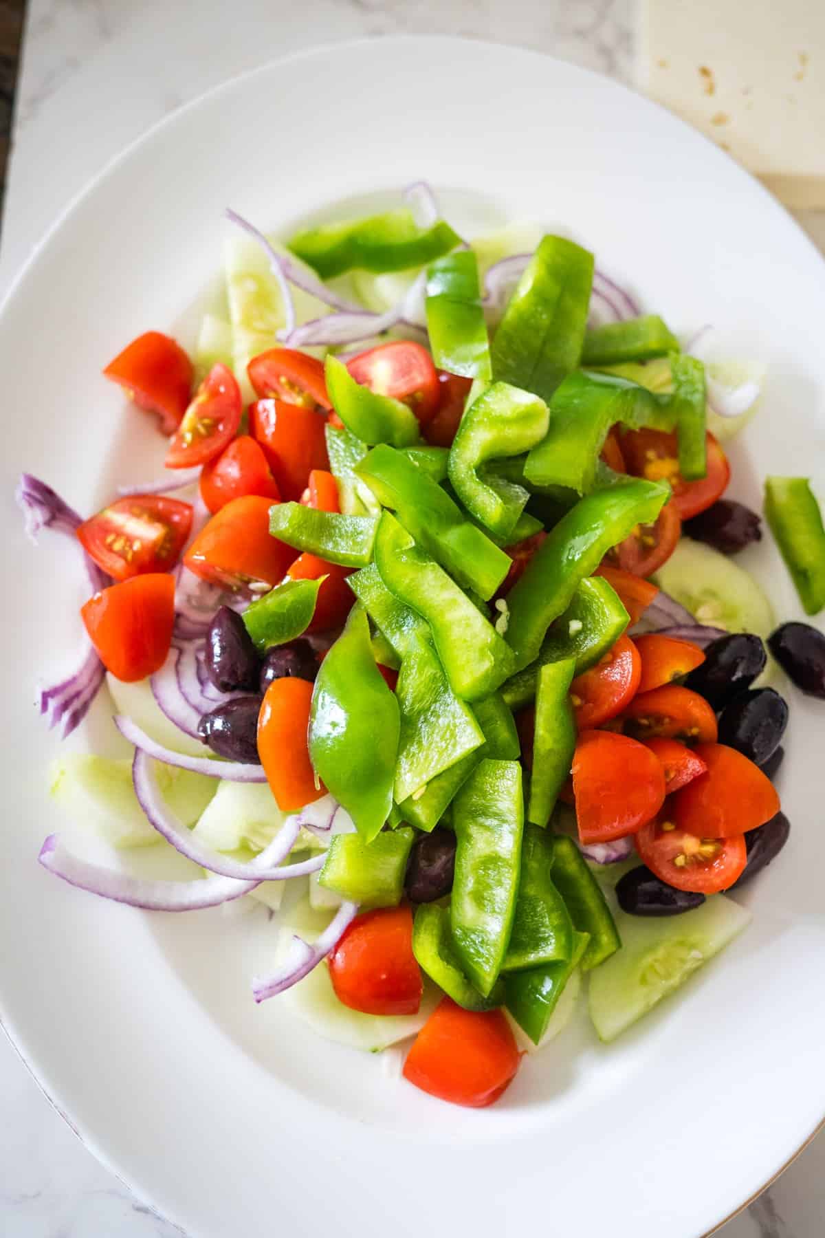 A white plate with a Greek village salad of sliced green bell peppers, red cherry tomatoes, black olives, red onion, and cucumber slices.