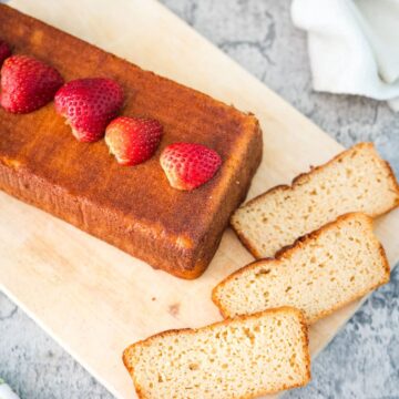 A loaf of pound cake topped with whole strawberries, partially sliced on a wooden cutting board with three slices laid out. A white cloth and stone countertop are visible in the background.