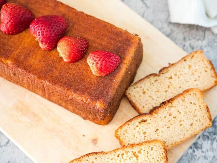 A loaf of pound cake topped with whole strawberries, partially sliced on a wooden cutting board with three slices laid out. A white cloth and stone countertop are visible in the background.