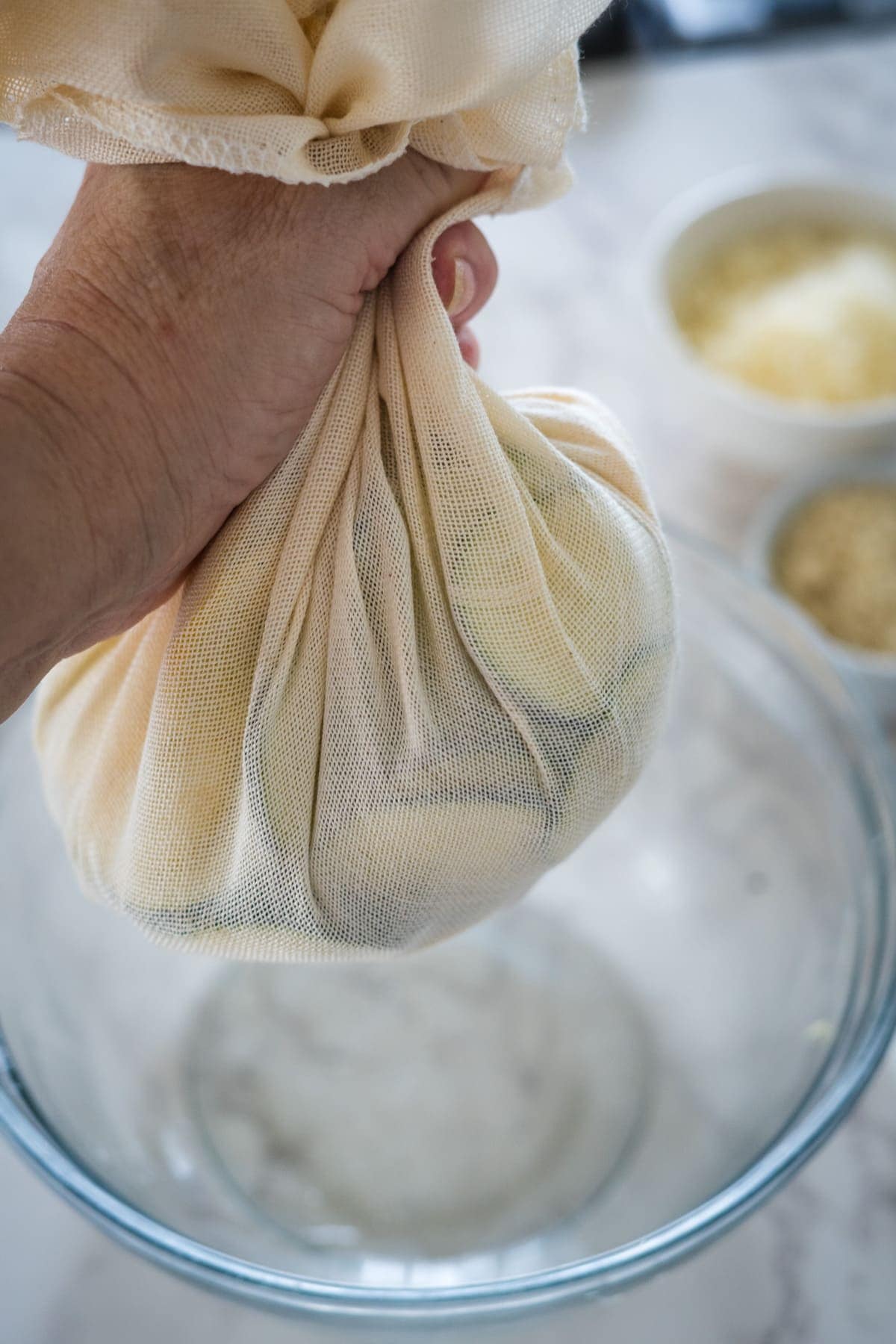 A hand is squeezing liquid from a bundle of shredded zucchini wrapped in cheesecloth over a glass bowl.